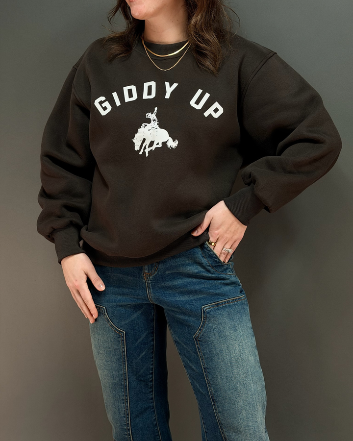 woman wearing brown crewneck sweatshirt that says giddy up with jeans in front of a gray background