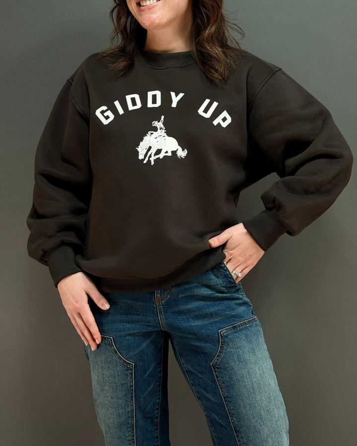 woman wearing brown crewneck sweatshirt that says giddy up with jeans in front of a gray background