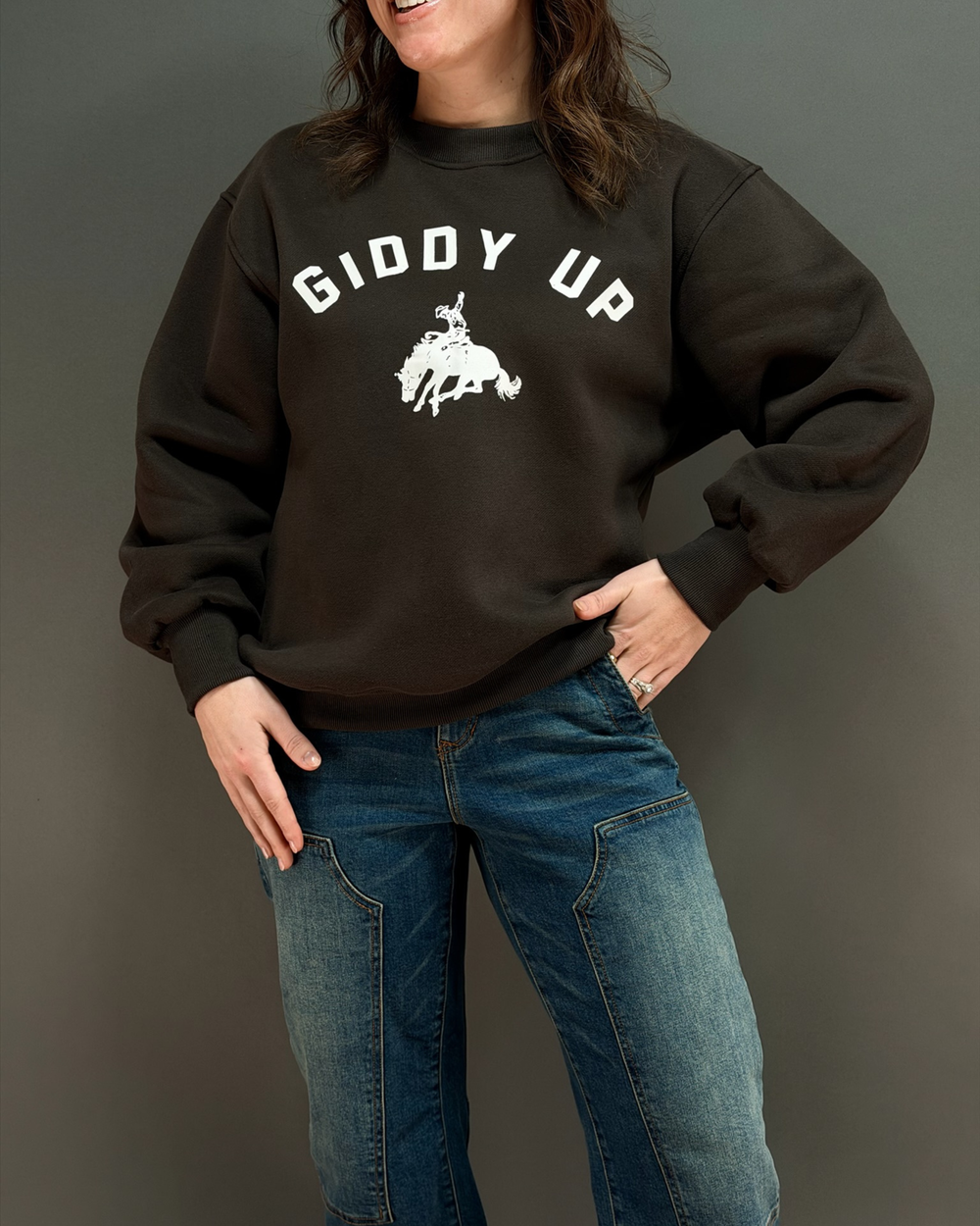 woman wearing brown crewneck sweatshirt that says giddy up with jeans in front of a gray background