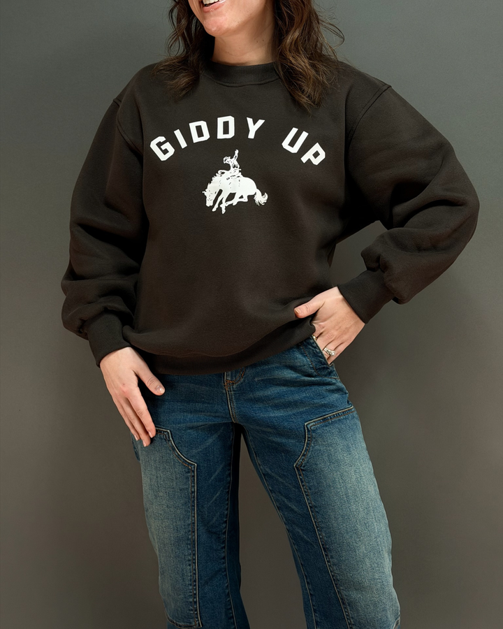 woman wearing brown crewneck sweatshirt that says giddy up with jeans in front of a gray background