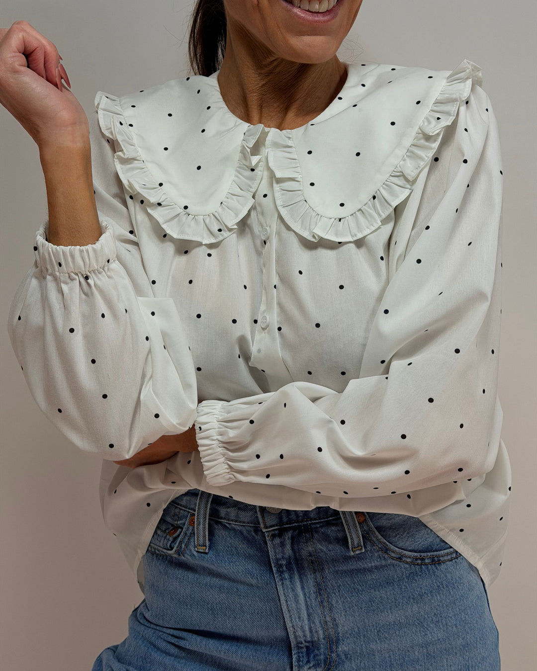 White blouse with black polka dots and ruffled collar worn by a person, against a neutral background.