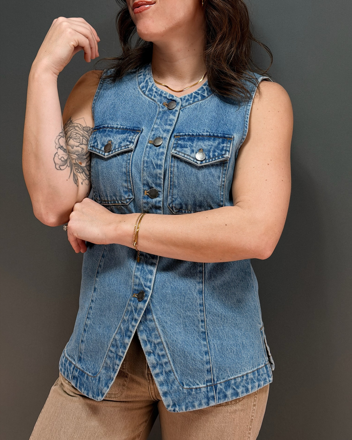 woman wearing a sleeveless denim vest  and brown pants against a gray background