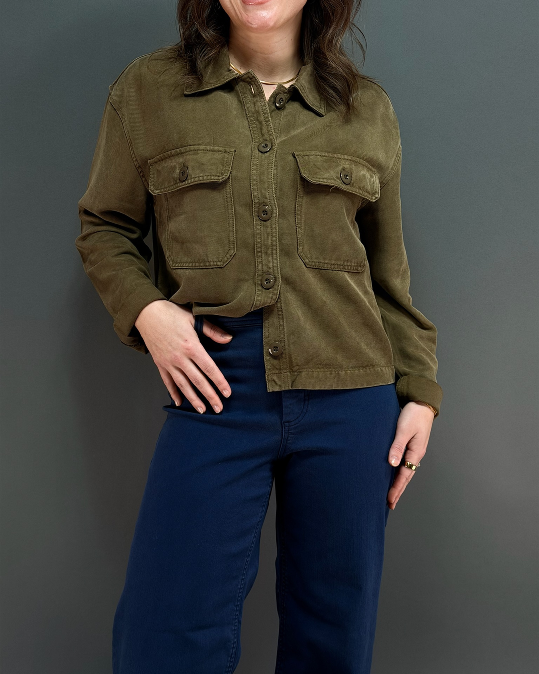 Woman wearing green button up utility style jacket with navy wide leg pants in front of a gray background