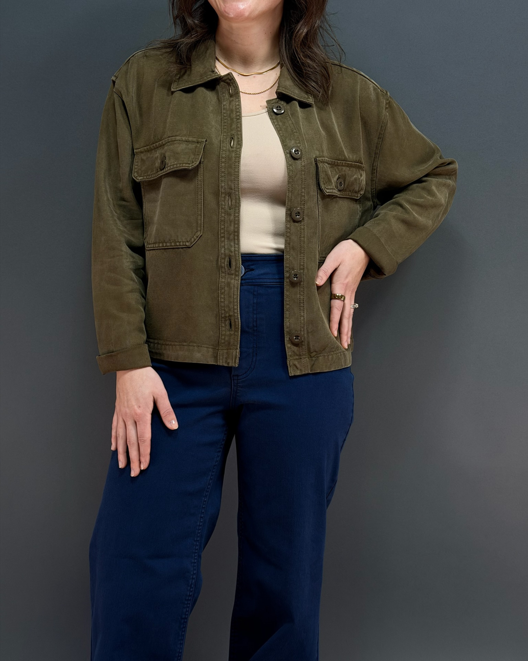 Woman wearing green button up utility style jacket with navy wide leg pants in front of a gray background