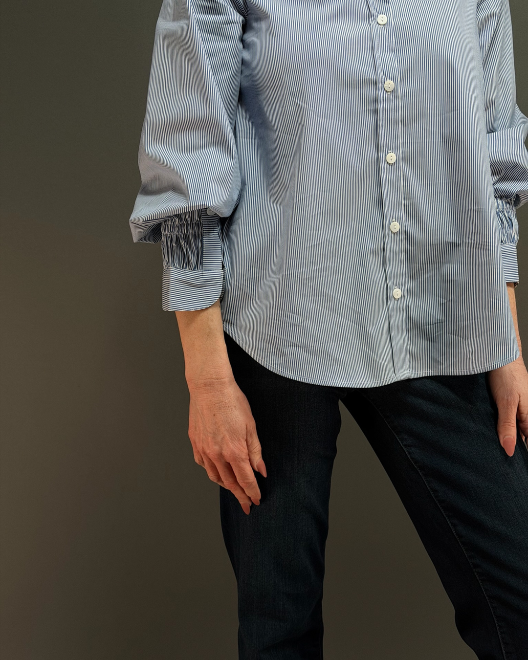 woman wearing a blue and white pinstripe button down top with ruched sleeves, paired with dark wash denim in front of a gray background