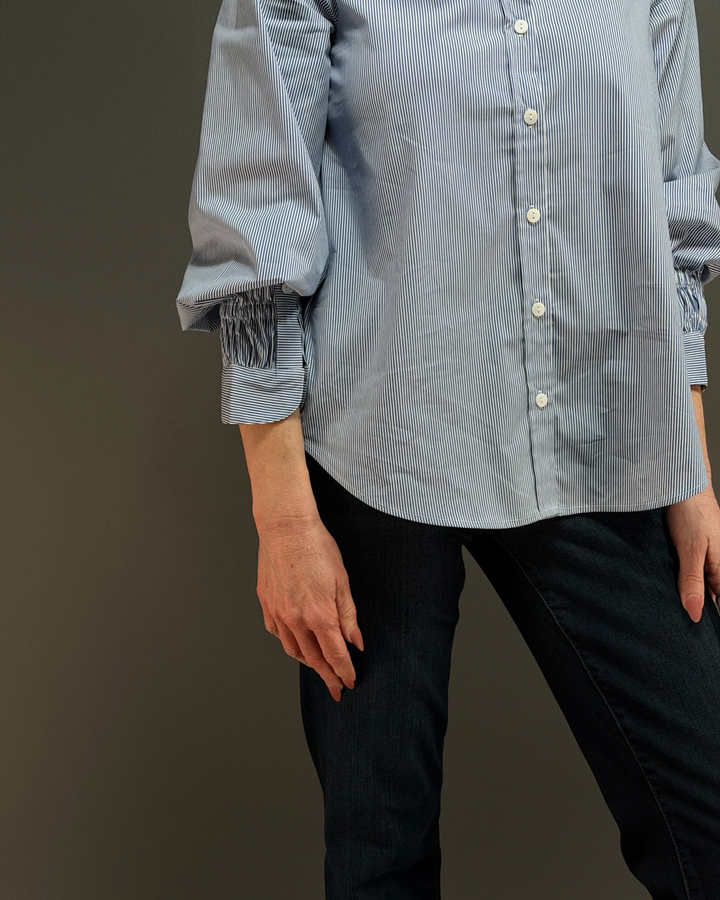 woman wearing a blue and white pinstripe button down top with ruched sleeves, paired with dark wash denim in front of a gray background