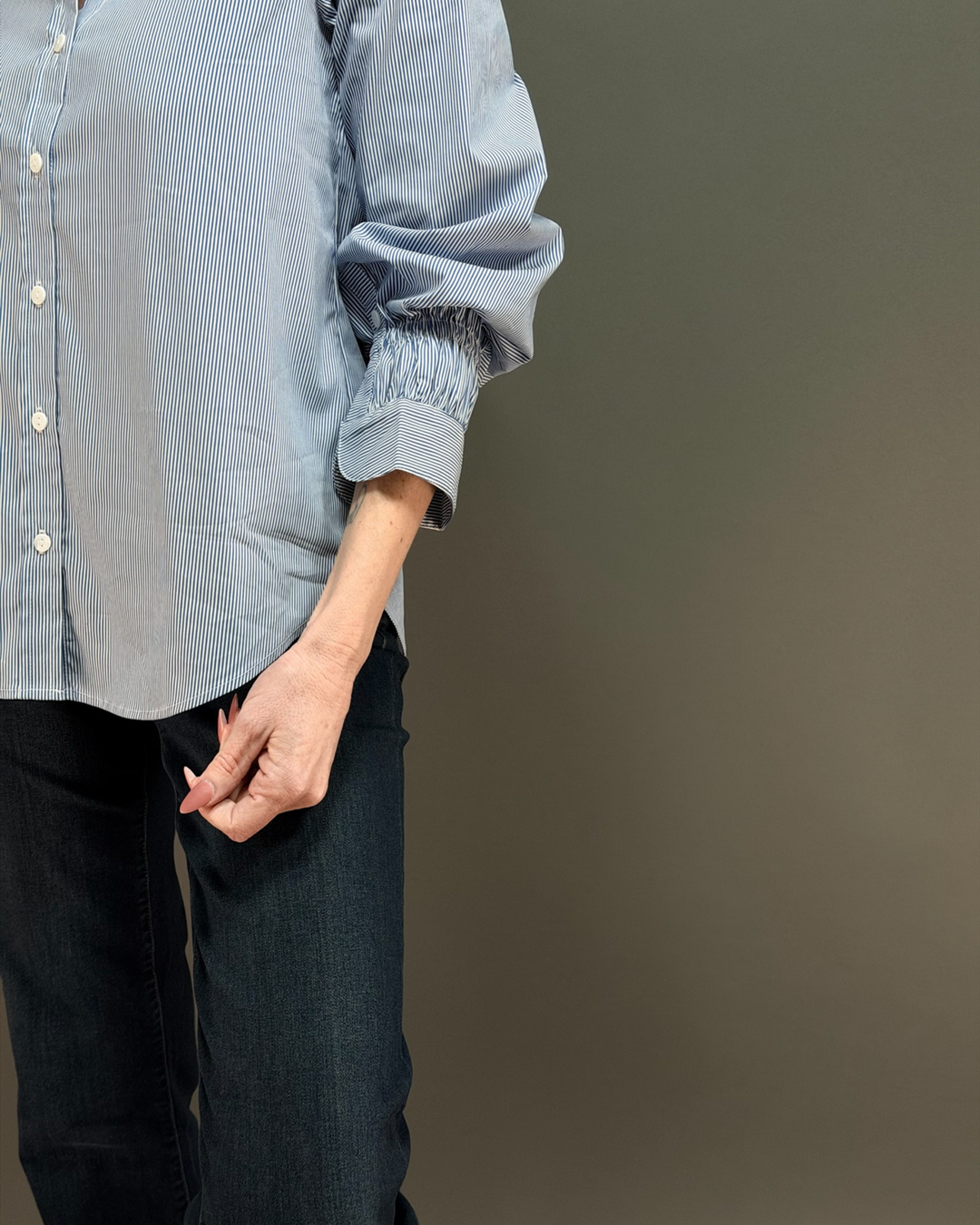 woman wearing a blue and white pinstripe button down top with ruched sleeves, paired with dark wash denim in front of a gray background