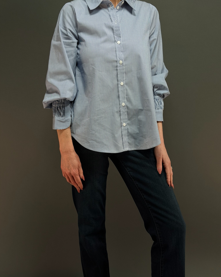 woman wearing a blue and white pinstripe button down top with ruched sleeves, paired with dark wash denim in front of a gray background