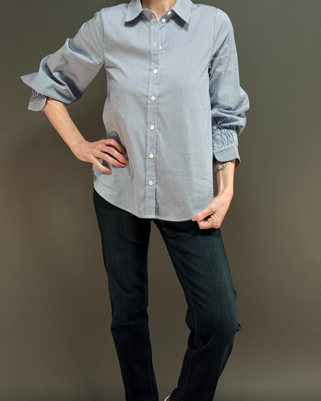woman wearing a blue and white pinstripe button down top with ruched sleeves, paired with dark wash denim in front of a gray background