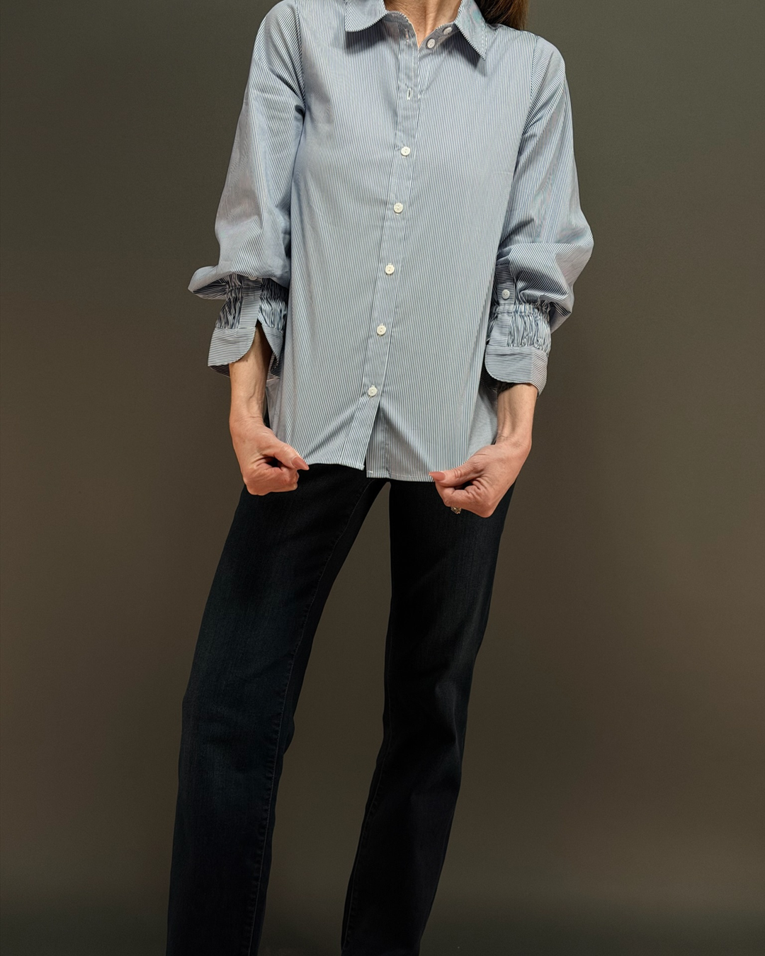 woman wearing a blue and white pinstripe button down top with ruched sleeves, paired with dark wash denim in front of a gray background