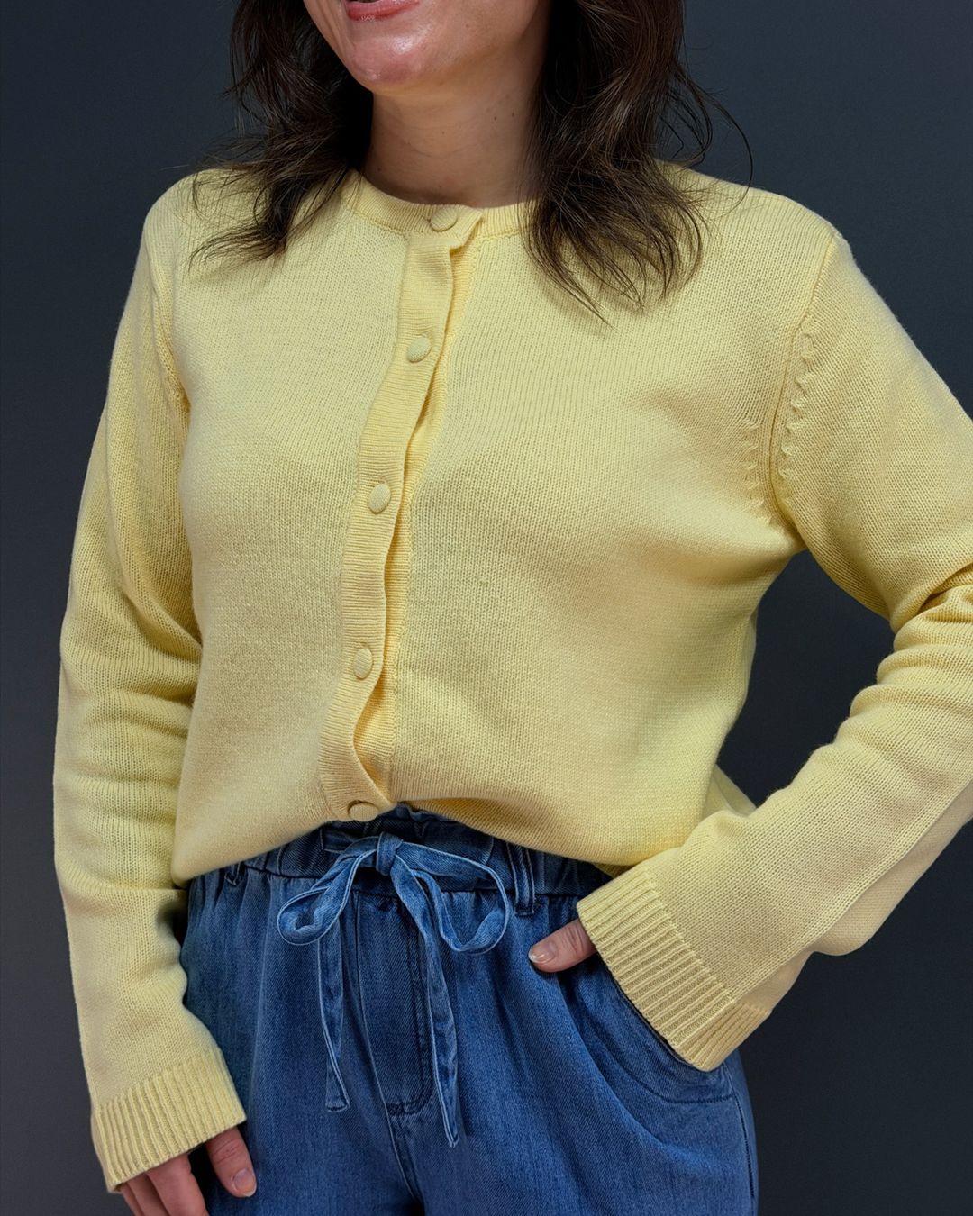 woman wearing yellow button down cardigan with blue tie waist pants in front of a gray background