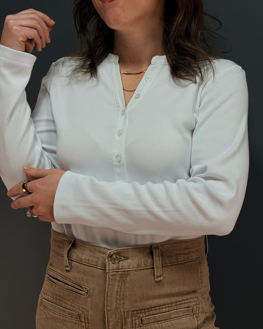 woman wearing white long sleeve top with brown pants in front of gray background