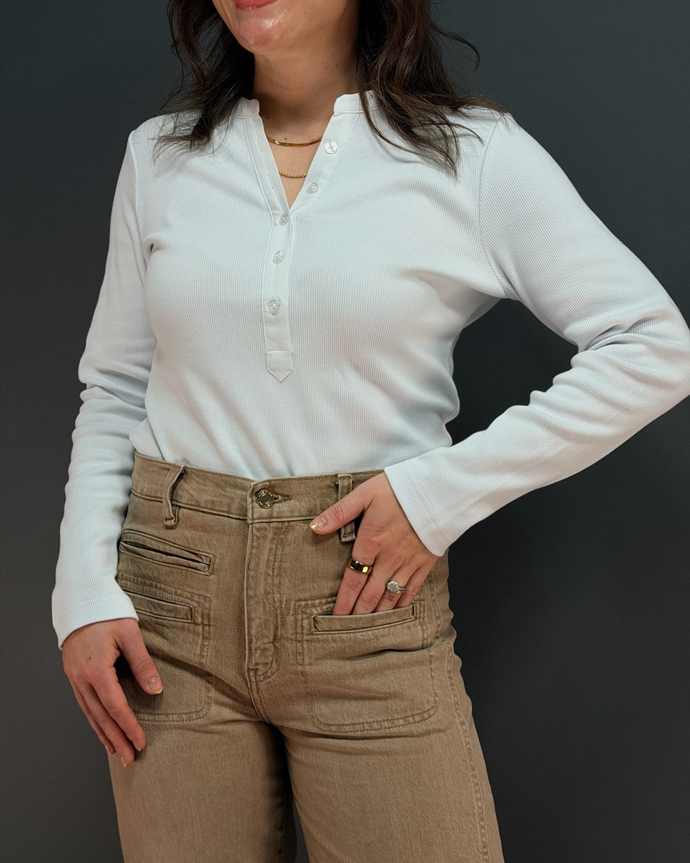woman wearing white long sleeve top with brown pants in front of gray background