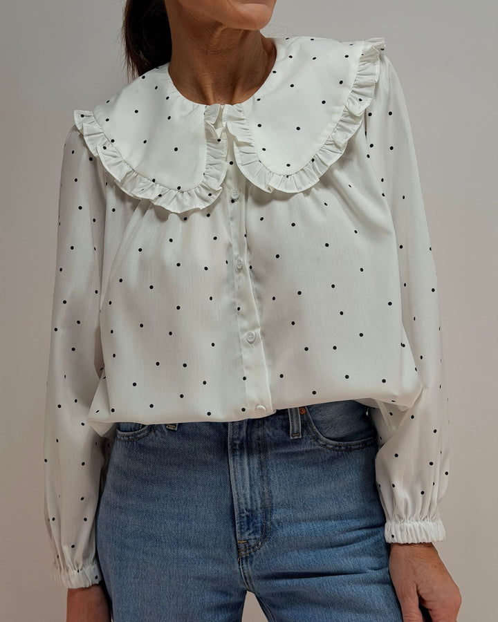 White blouse with black polka dots and ruffled collar worn by a person, against a neutral background.