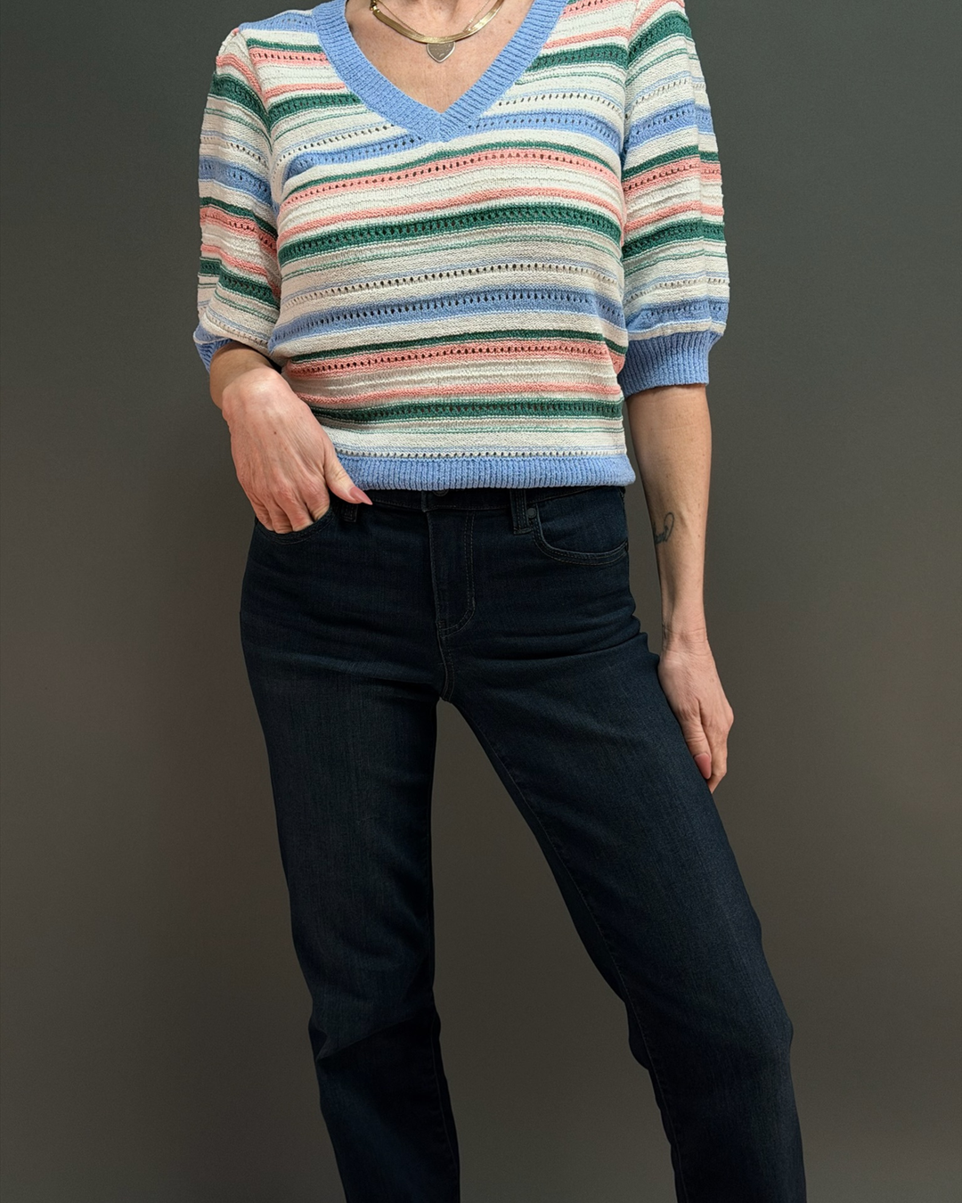 woman wearing a short colorful striped knit sweater with dark wash jeans in front of a gray background