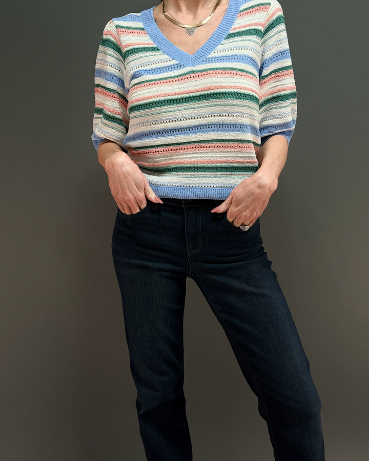 woman wearing a short colorful striped knit sweater with dark wash jeans in front of a gray background