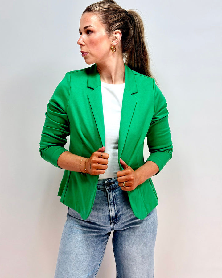Woman wearing a green blazer over a white shirt and blue jeans on a light gray background