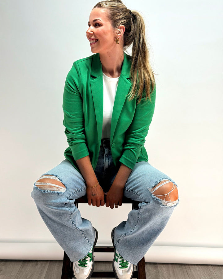 Woman wearing a green blazer, white shirt, and ripped jeans sitting on a chair.