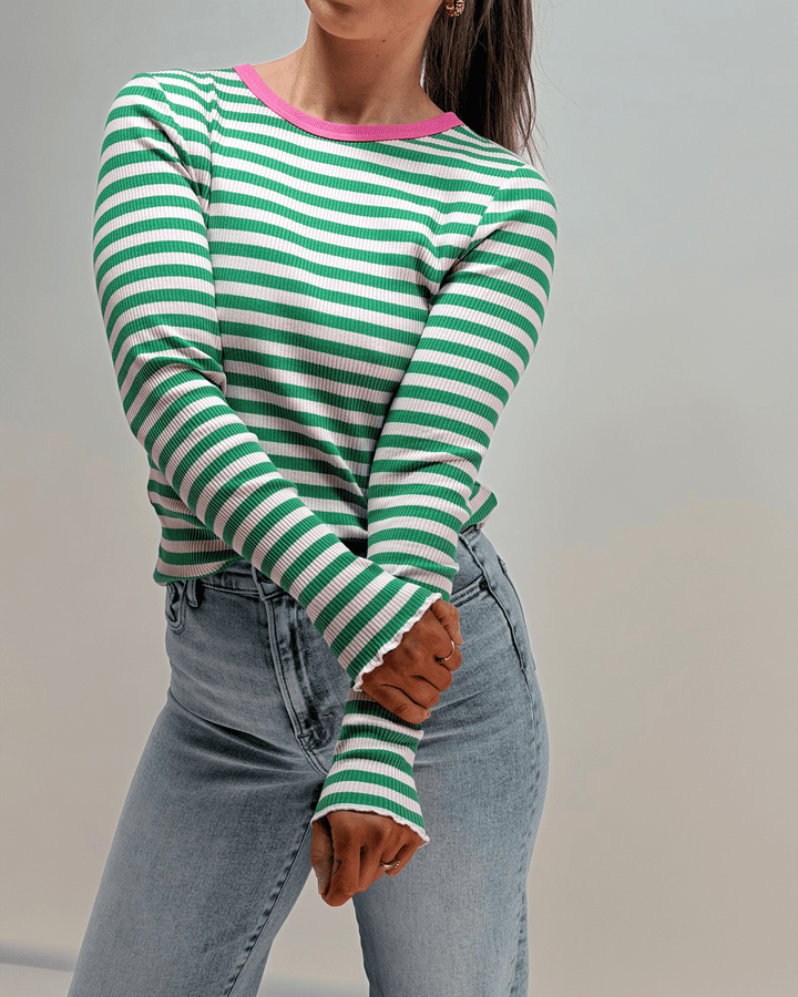 Person wearing a green and white striped sweater with pink trim, paired with light blue jeans against a neutral background.