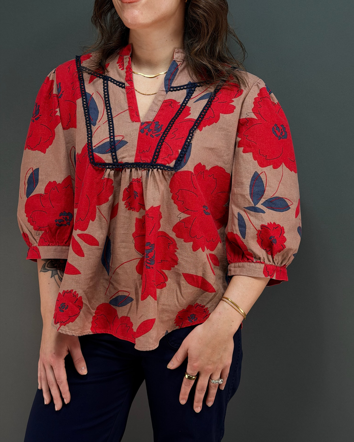 woman wearing tan and red floral blouse with navy pants in front of a gray background